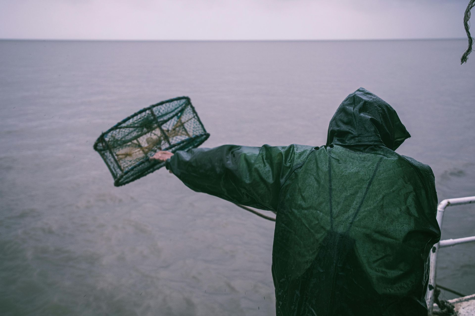 Person in green rain gear holding a crab trap on a boat, overcast water in background.