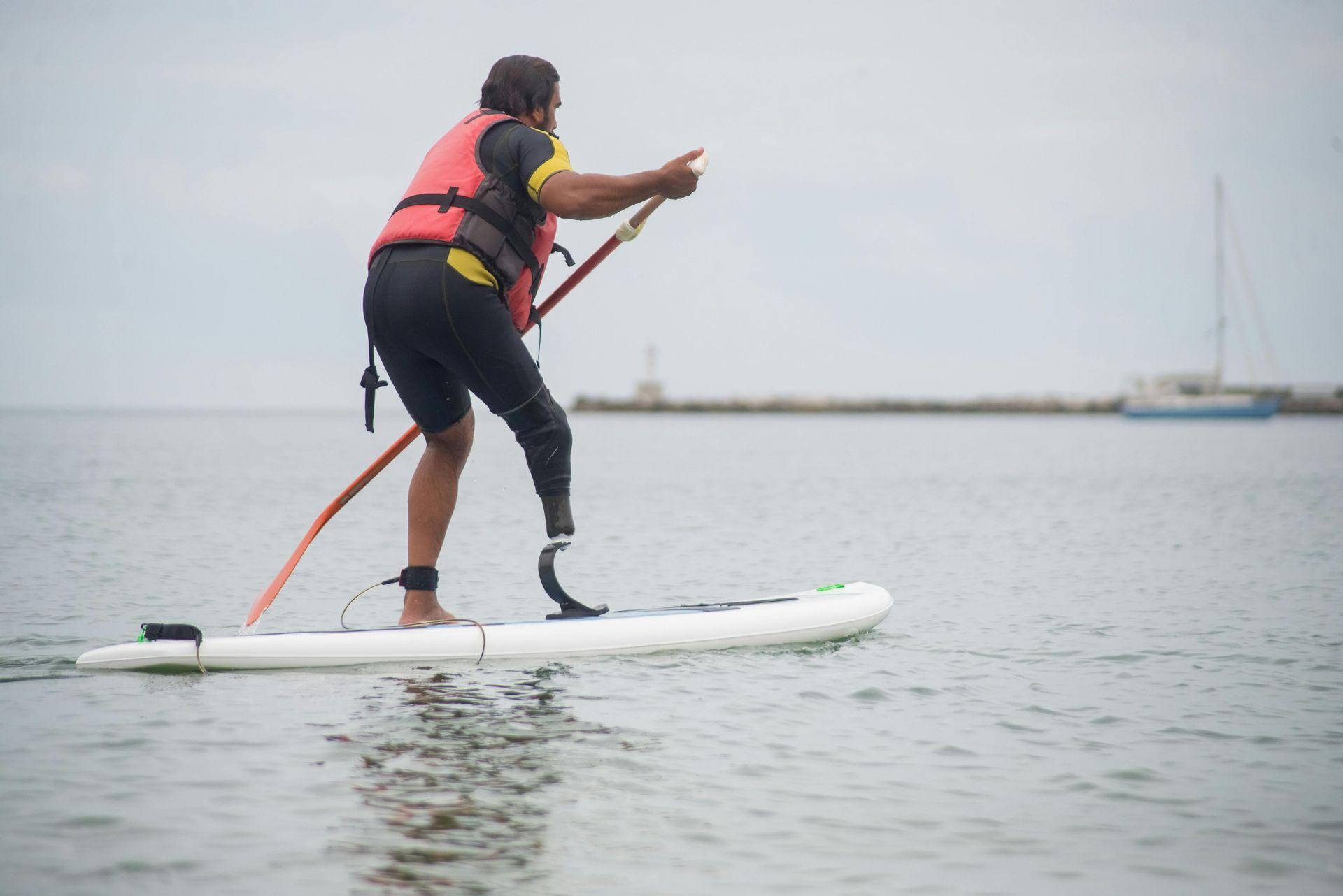Man with prosthetic leg paddleboards in calm water, wearing a life vest.