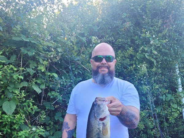 Man with a beard and sunglasses holds up a fish near green foliage in sunlight.