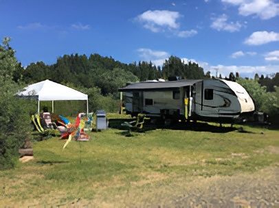 RV parked at a campsite with a white canopy, chairs, and surrounding trees under a blue sky.