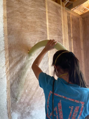 A person in a blue t-shirt and mask holds a flexible hose, blowing loose-fill insulation into a wall cavity.