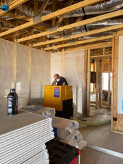 A worker in a mask stands behind a yellow machine in a construction site with stacks of drywall in the foreground.
