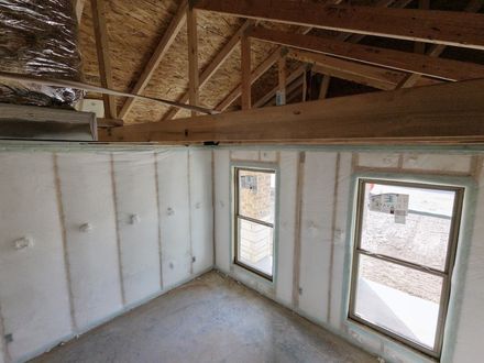 An interior view of a room under construction with insulated walls, two windows, and an exposed wooden roof truss system.