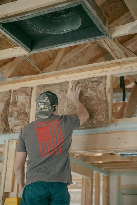 A person in a gray graphic t-shirt and camouflage hat installs brown insulation between wooden wall studs.