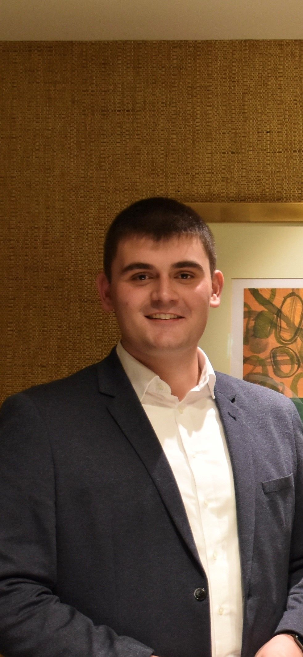 Ein Mann in einem dunkelblauen Blazer und weißem Hemd lächelt, er steht drinnen vor einer strukturierten Wand.