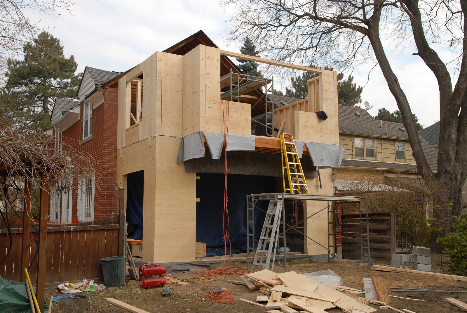Building under construction with wood framing attached to a red brick house; scaffolding and ladder visible.