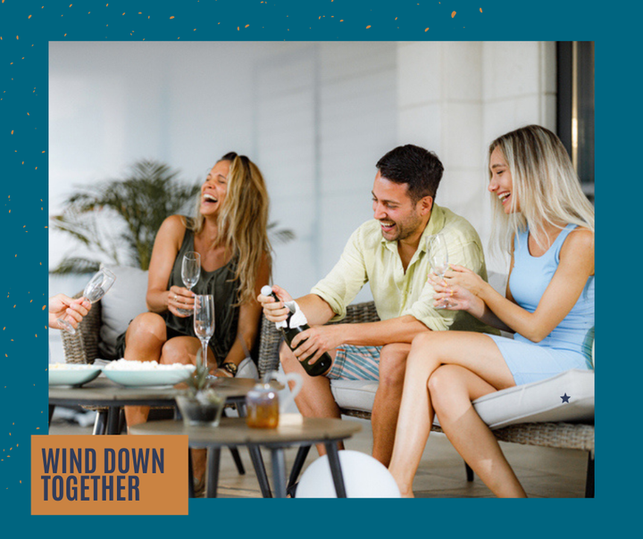 Three friends laugh while opening champagne on a patio.