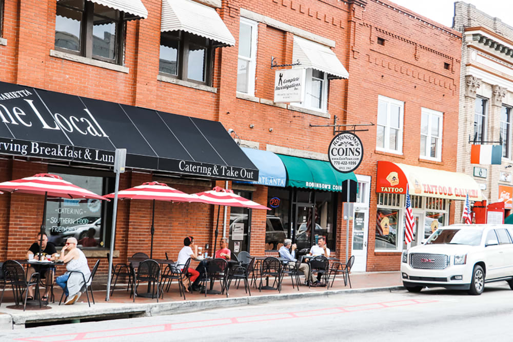 Street scene with brick buildings, shops, and outdoor seating under red and white umbrellas. People are dining and a white SUV is parked on the street.