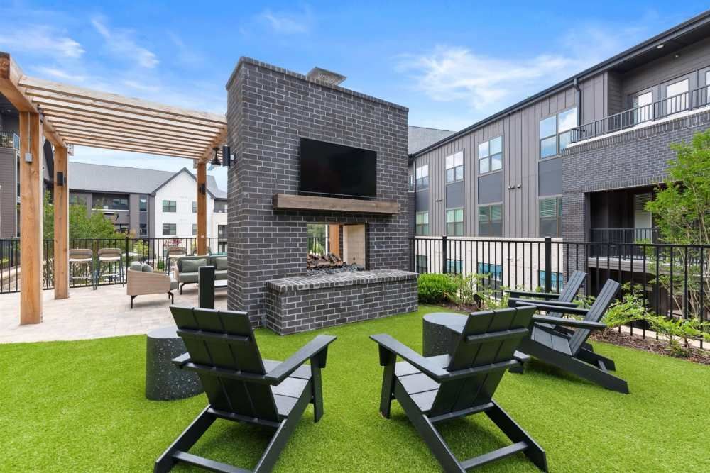 Outdoor lounge area with fireplace, TV, and seating. Artificial turf, dark brick fireplace, and modern black chairs under a blue sky.