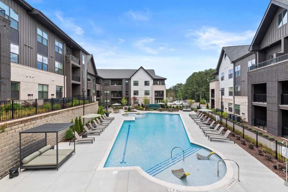 Apartment complex with a pool, featuring gray buildings, lounge chairs, and a cabana on a sunny day.