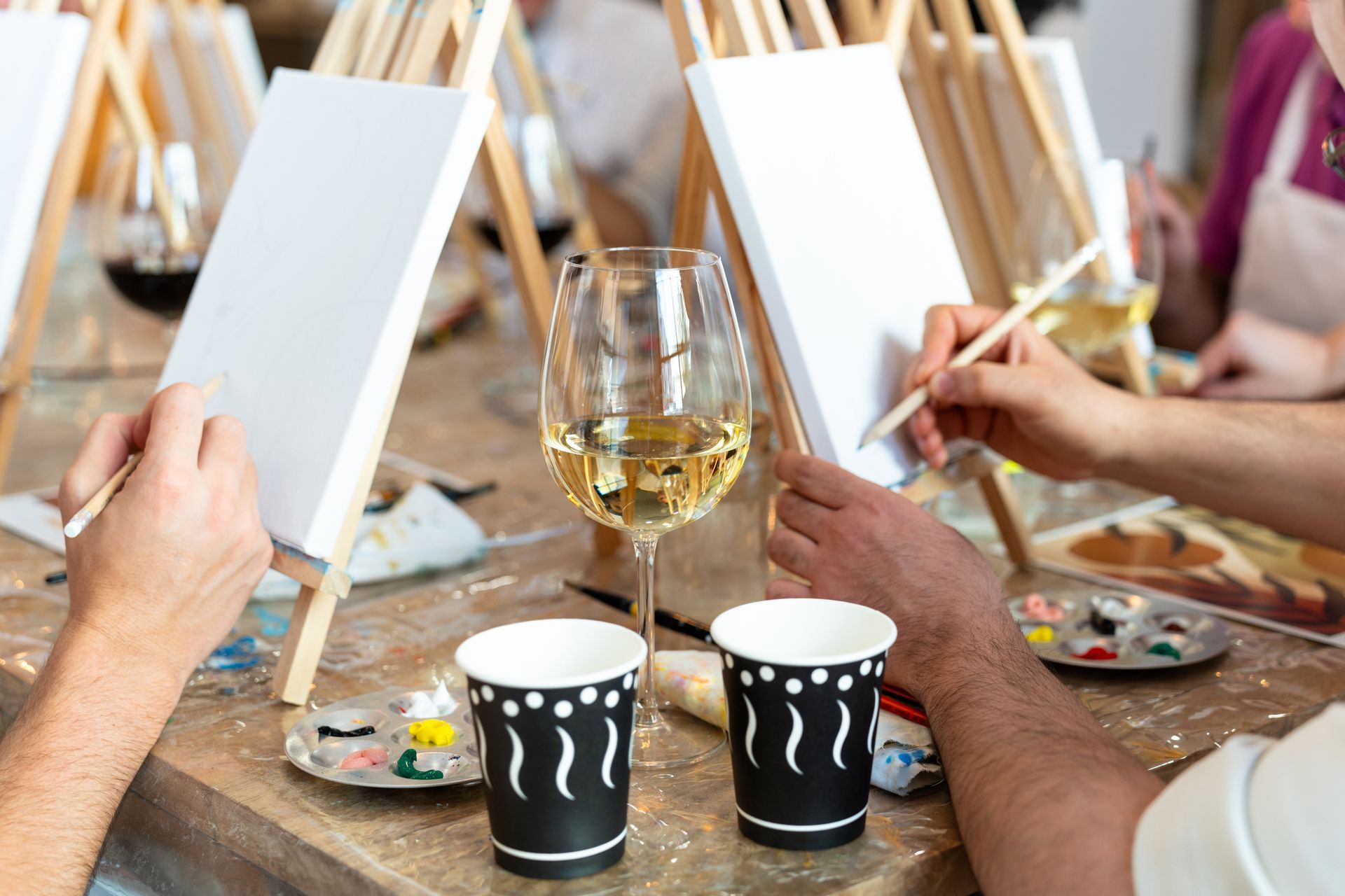 People painting on easels at a paint and sip event. Wine glass and cups on the table.