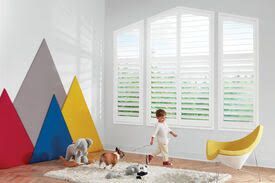 A child is playing with a dog in a room with shutters.