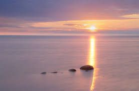 The sun is setting over the ocean with rocks in the foreground.