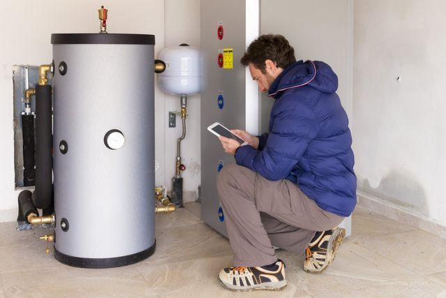 A man is kneeling down in front of a heater and looking at a tablet.