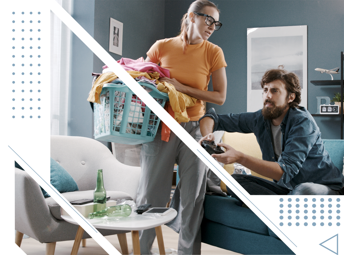 A man is playing a video game while a woman is holding a laundry basket.