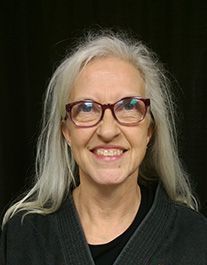 A woman with long gray hair and glasses is smiling for the camera.