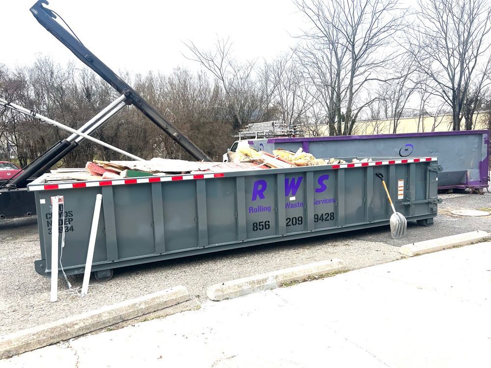 Gray RWS dumpster with debris, parked in front of a purple dumpster. Broom leaning on the side.
