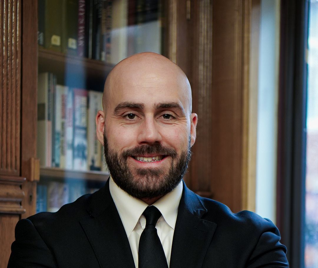 A man in a suit and tie is standing with his arms crossed in front of a bookshelf.