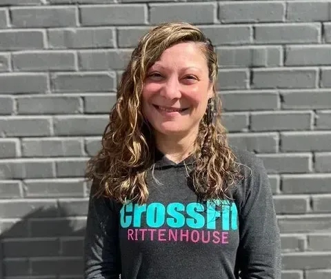 A woman wearing a crossfit shirt is smiling in front of a brick wall.