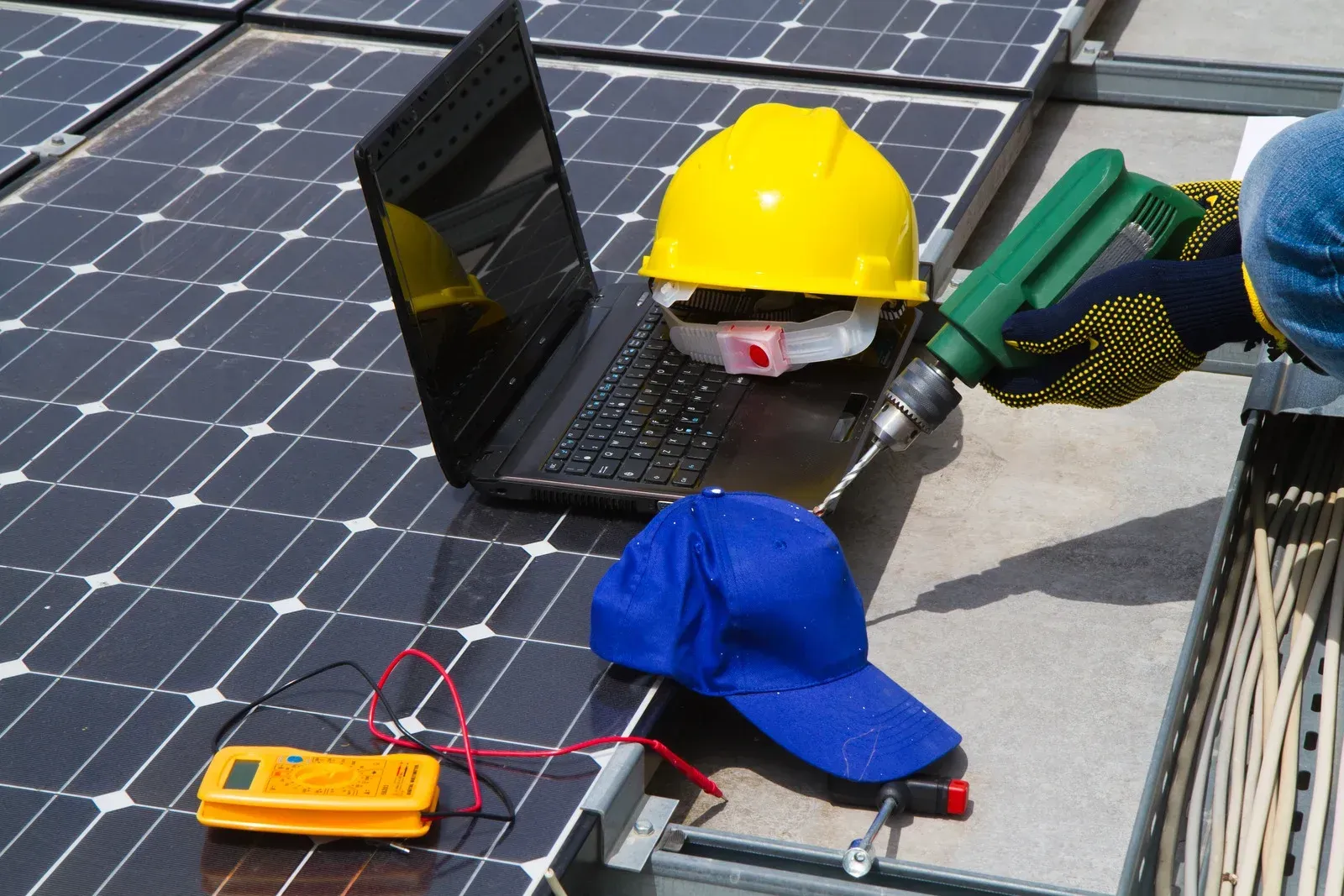 Person using a drill near solar panels, laptop, yellow hard hat, and voltage meter on a rooftop.
