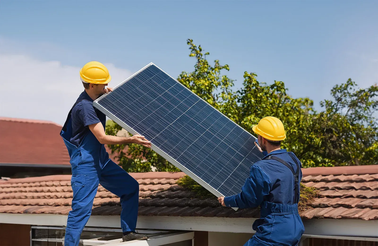 Two workers in blue overalls and yellow hard hats carry a solar panel on a rooftop.