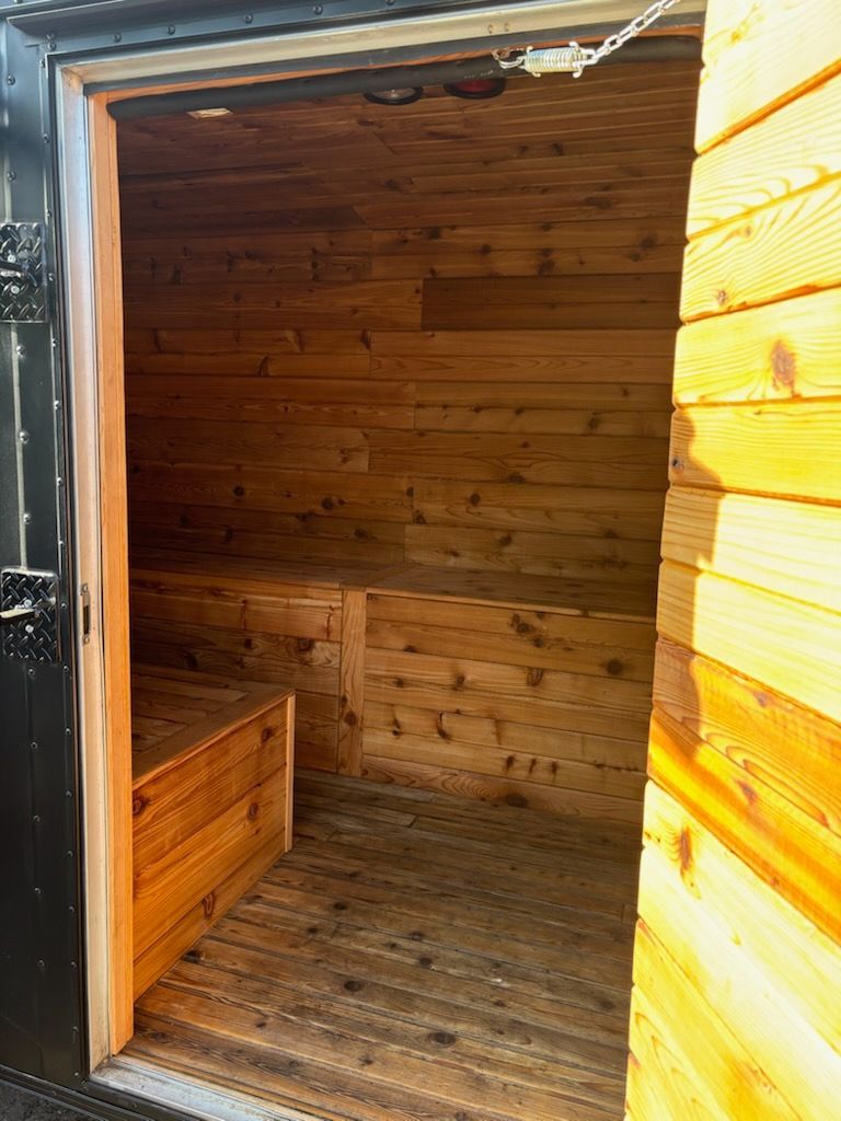 Interior of a wooden sauna with benches and a wooden floor. The door is open.