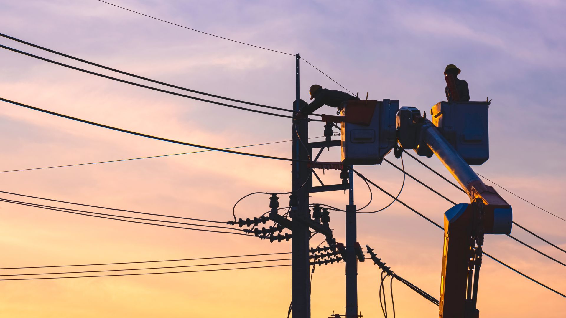 A silhouette of two electricians performing bucket truck service on an electrical line at sunset  