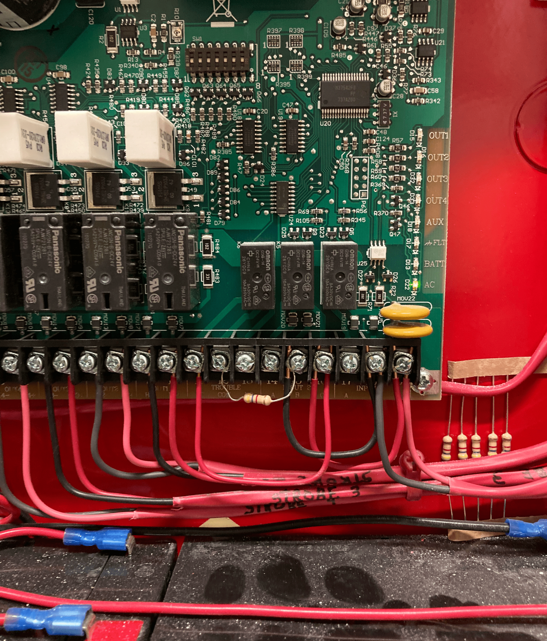 Fire alarm control panel's circuit board with wires. Red, black wires connected to the board. Red metal enclosure.