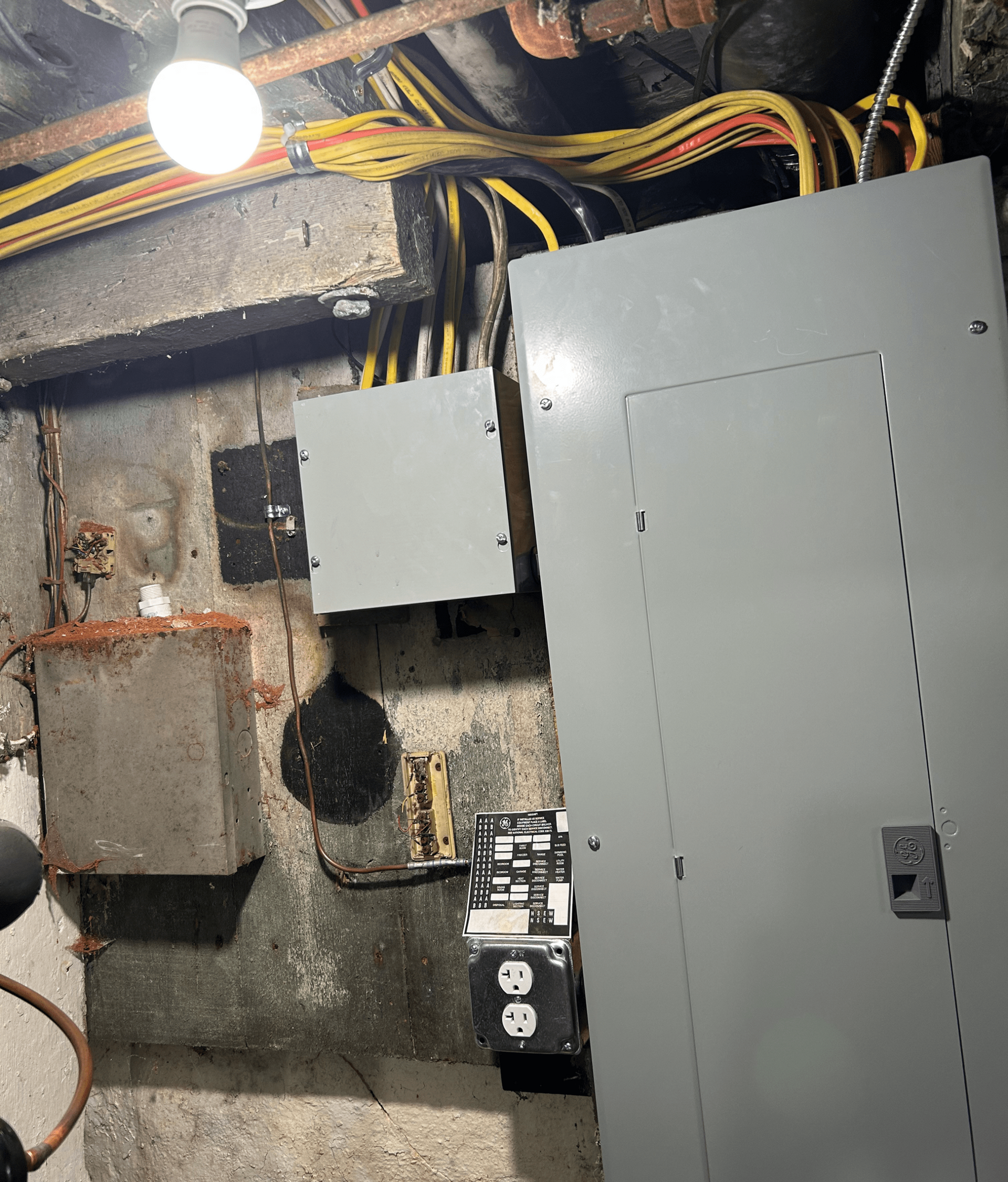 Electrical panels and wiring in a concrete basement. Gray panels, exposed wires, and a lightbulb illuminate the area.
