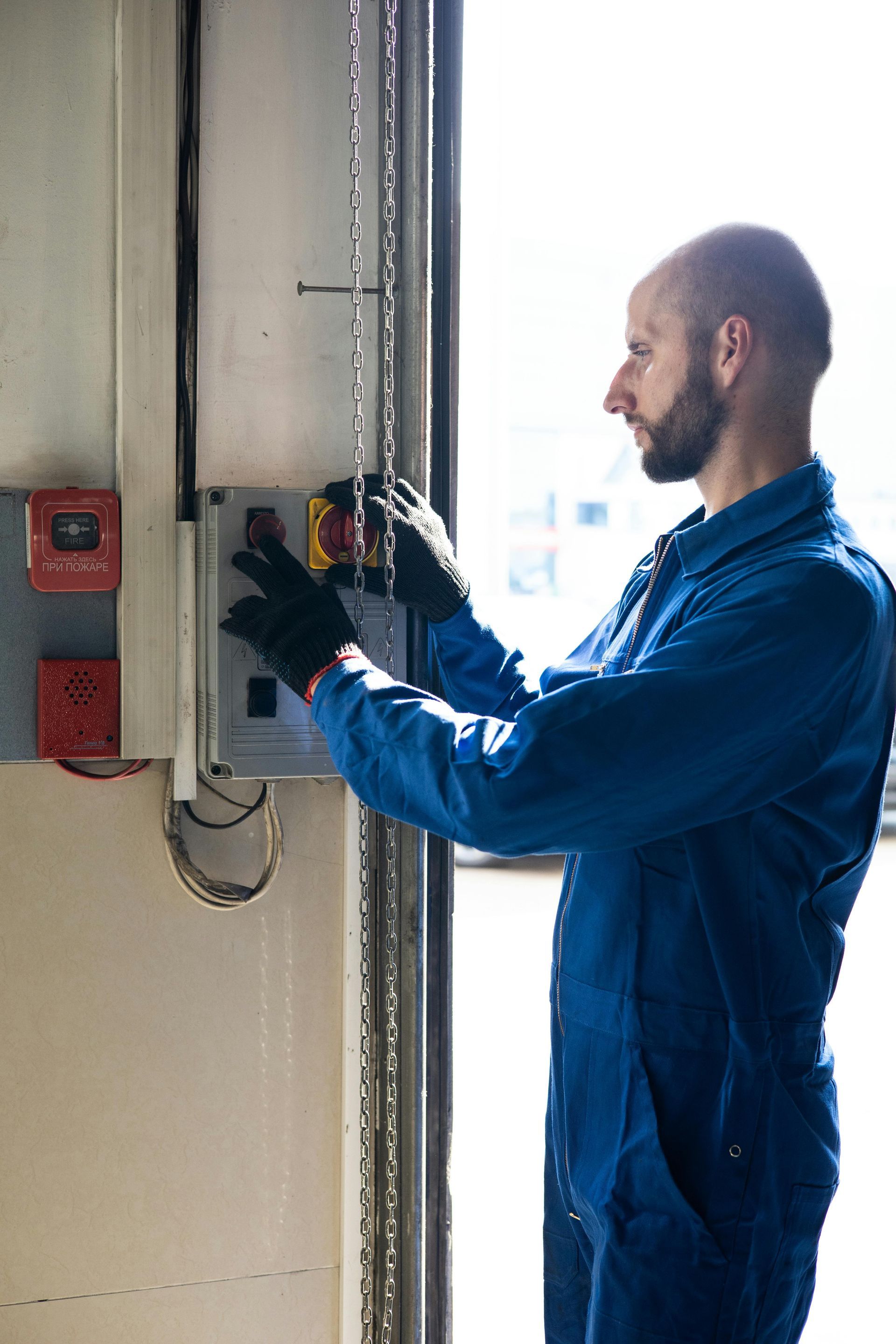 Man in blue overalls operating a control panel near an open door; wearing black gloves.