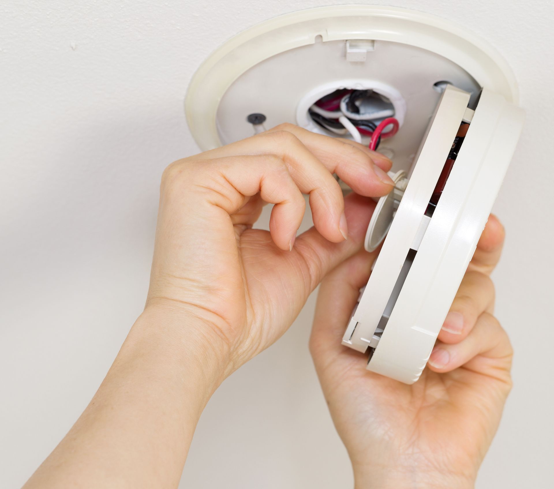 Hands installing a white smoke detector on a white ceiling. Wires visible.