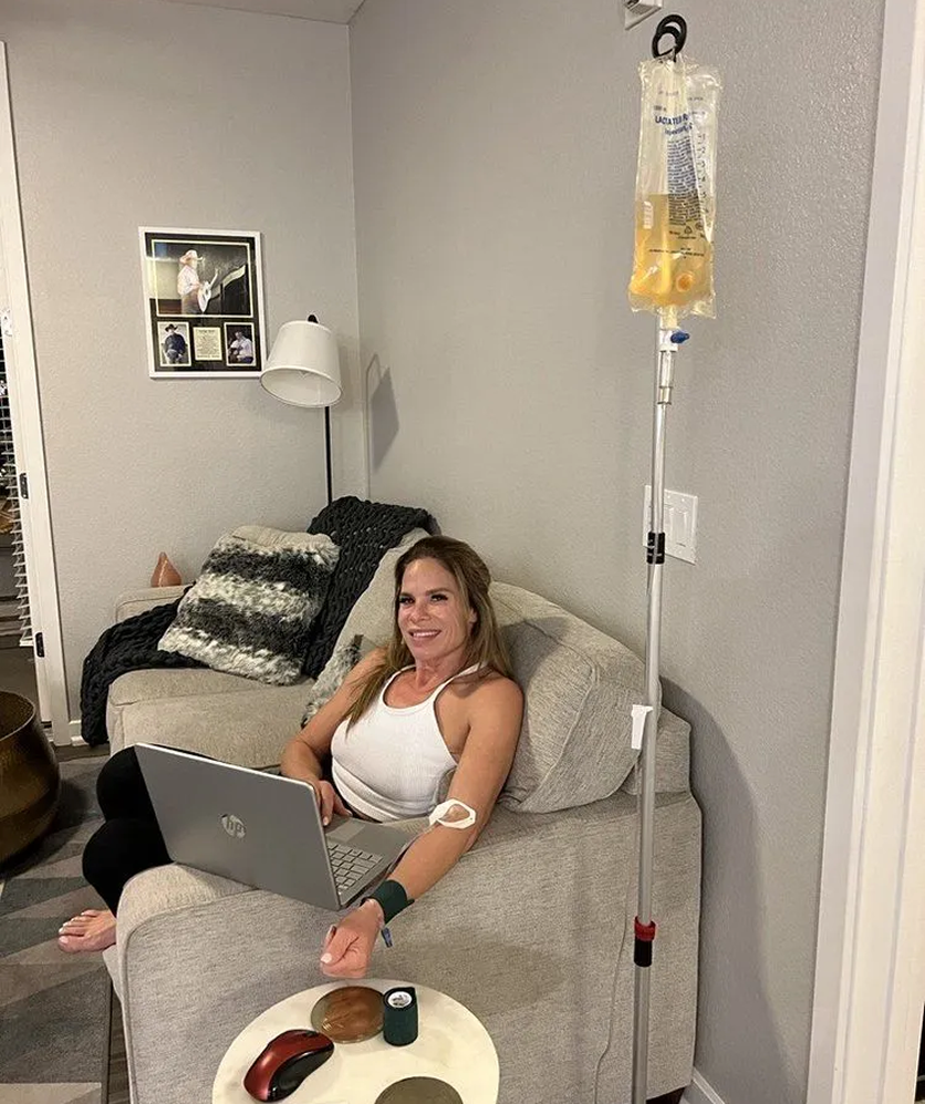 A woman is sitting on a couch with a laptop and an iv bag.