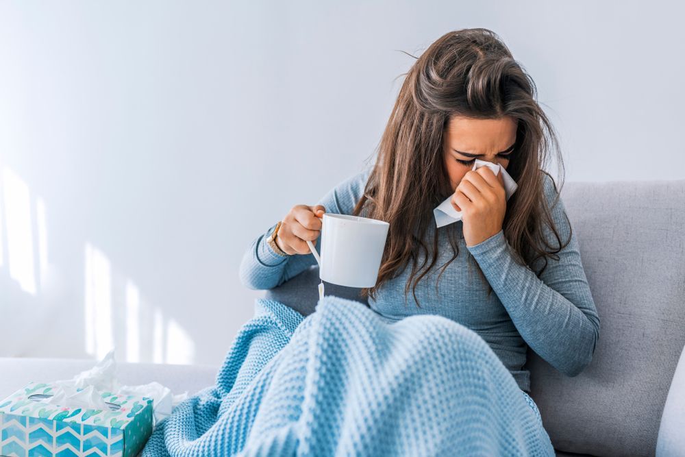 A woman is sitting on a couch holding a cup of coffee and blowing her nose.