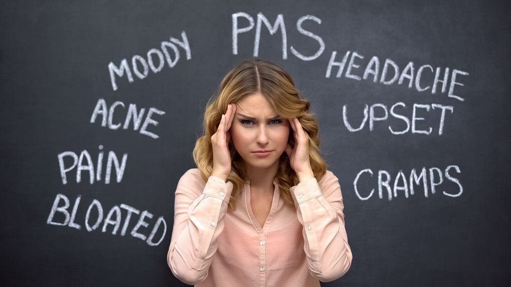 A woman is holding her head in front of a blackboard with words written on it.