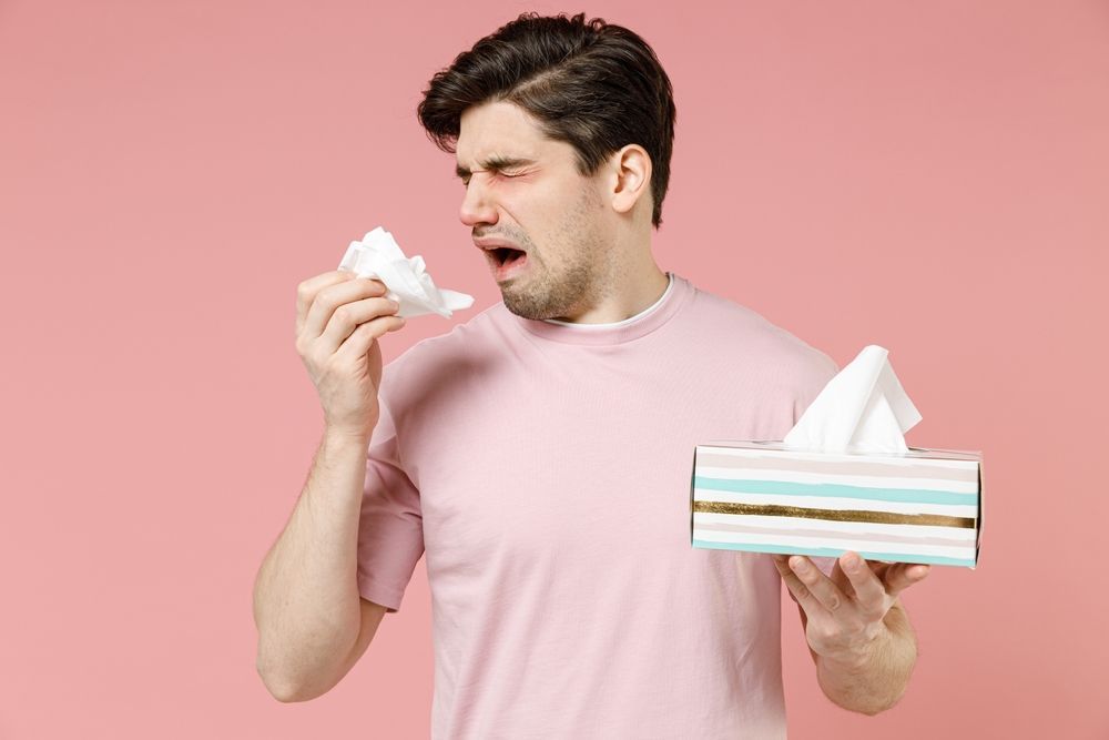 A man is blowing his nose into a napkin while holding a box of tissues.