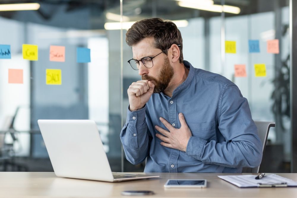 A man is coughing while sitting at a desk in front of a laptop computer.