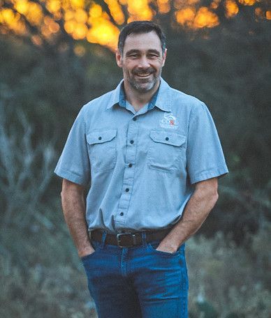 A man in a blue shirt and jeans is standing in a field with his hands in his pockets.