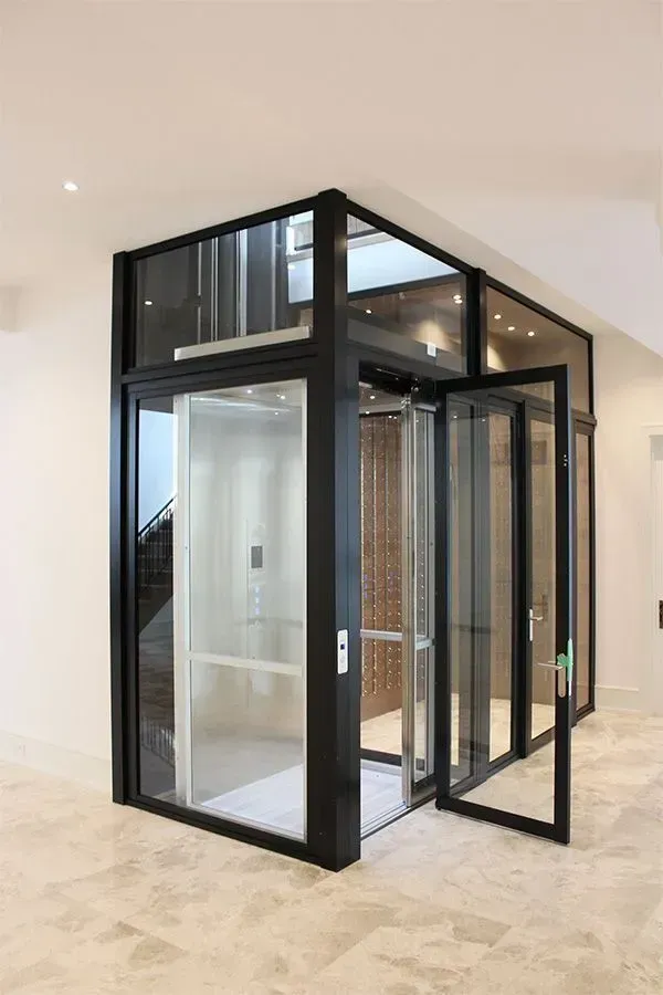 Modern home elevator with glass walls and black framing, in a light-filled interior.