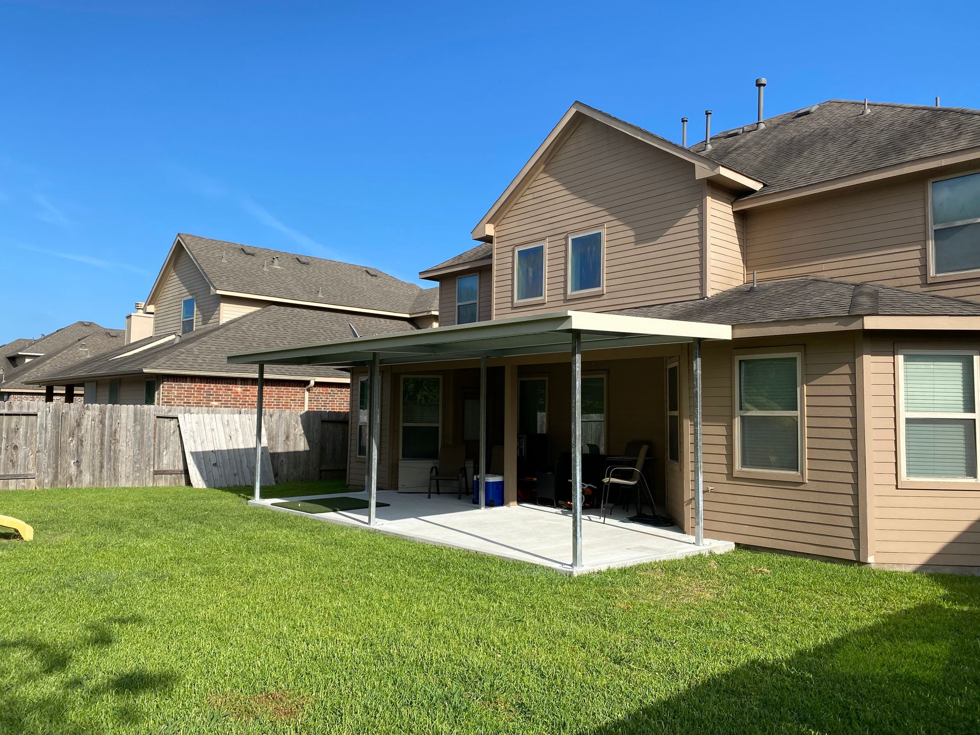 Backyard with a patio cover, green grass, and a two-story house under a blue sky.
