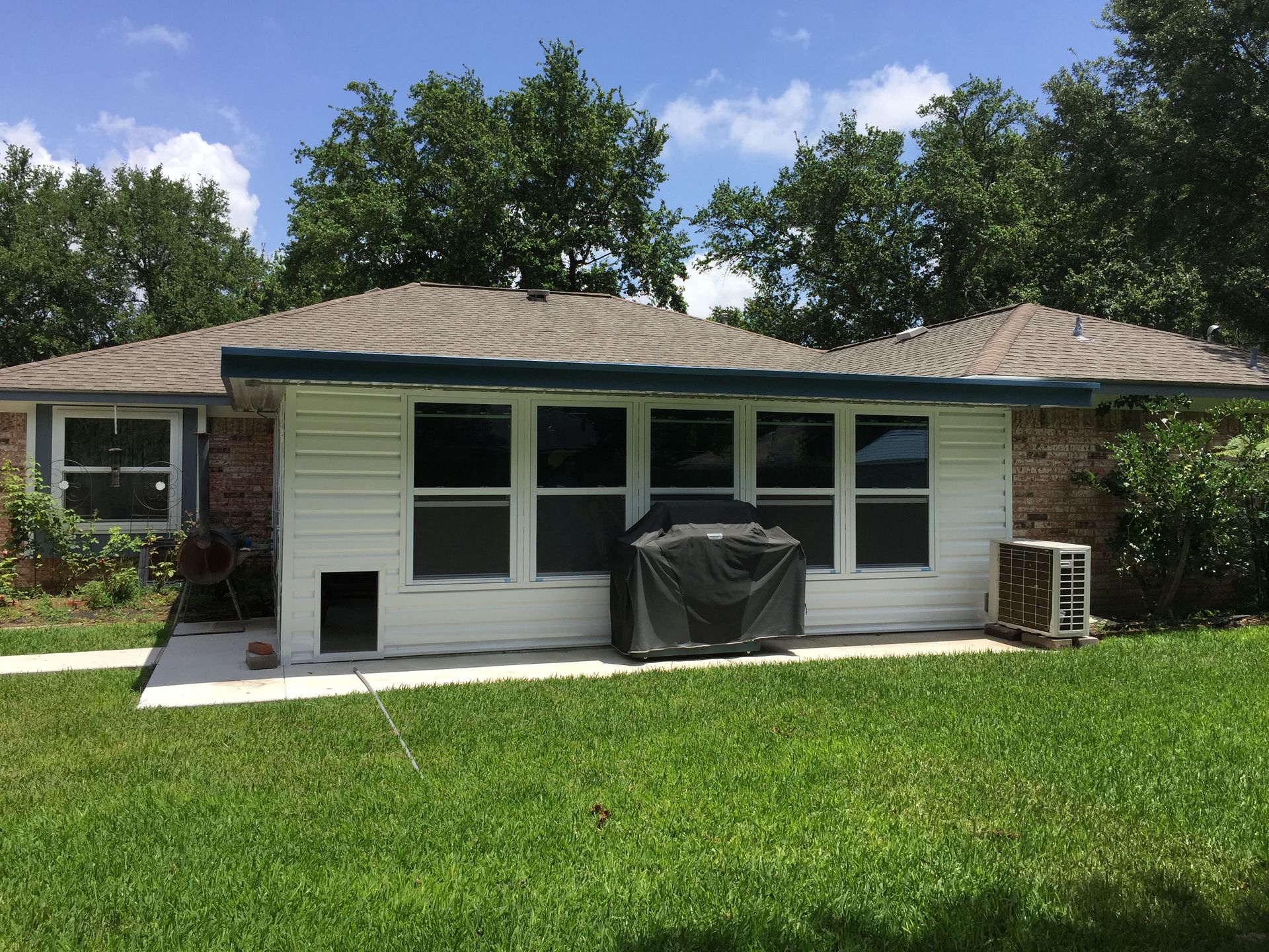 White-sided building with windows, a covered grill, and air conditioner, in a grassy backyard, under a sunny sky.