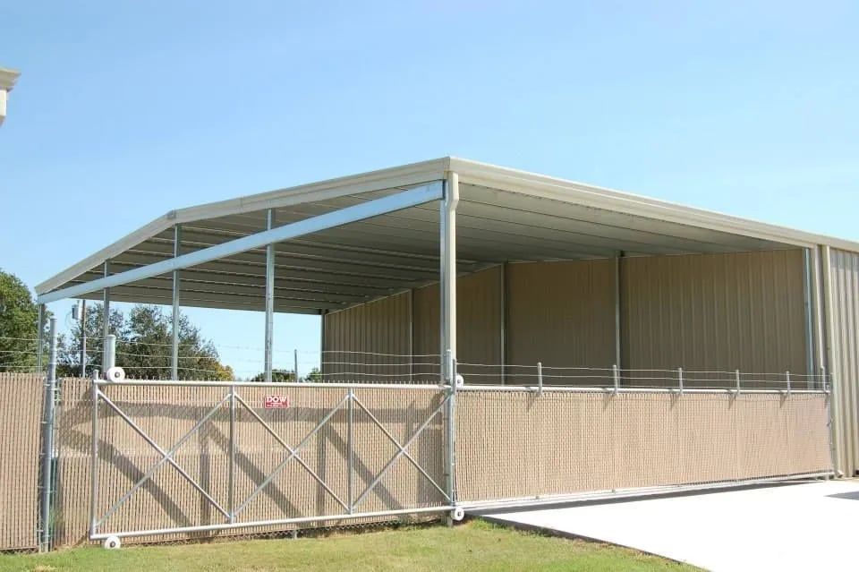 Open-air horse shelter with a tan roof and walls, in a grassy area with a clear sky.