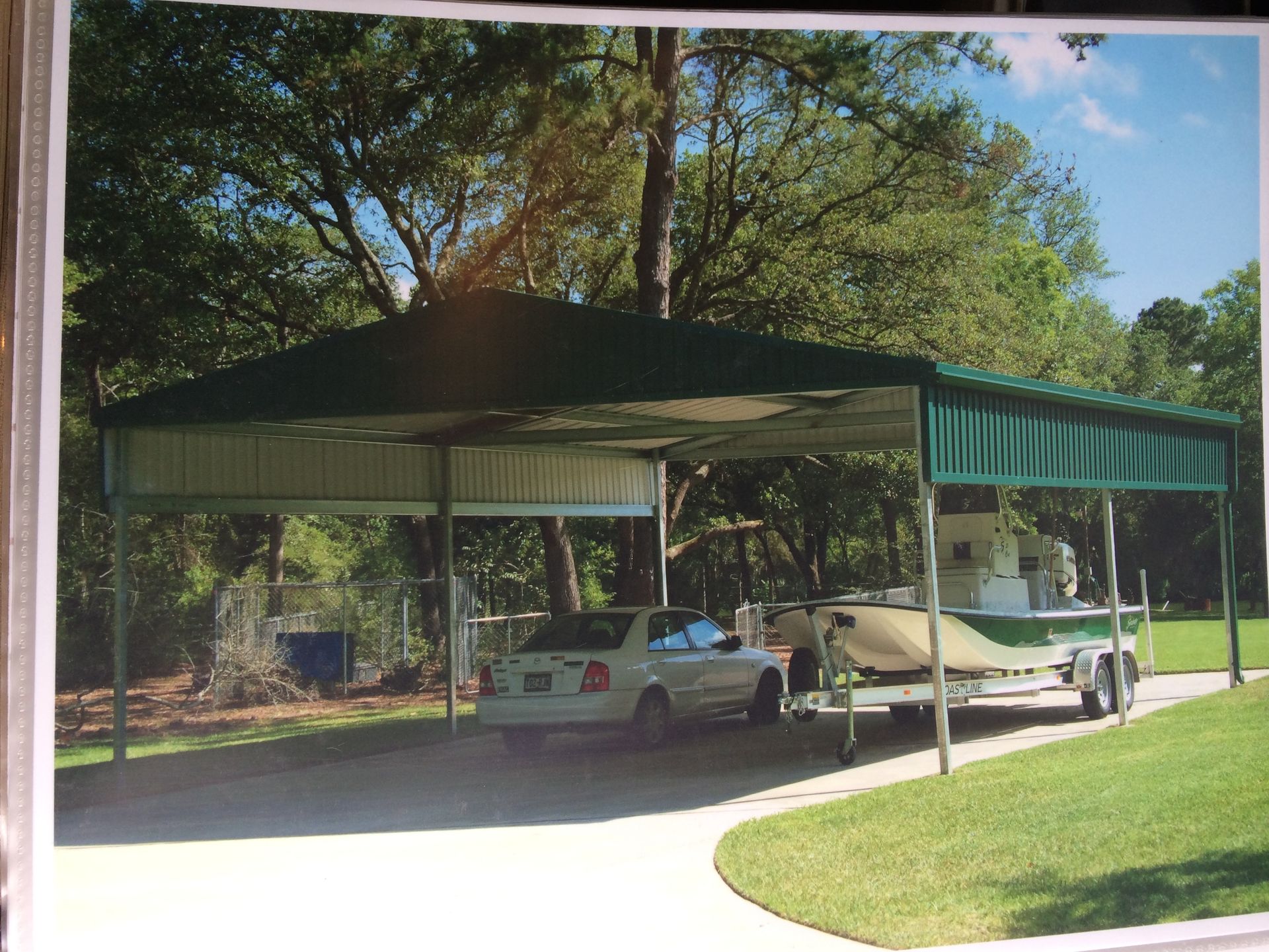 Green carport sheltering a white car and boat trailer on a driveway, with trees in the background.