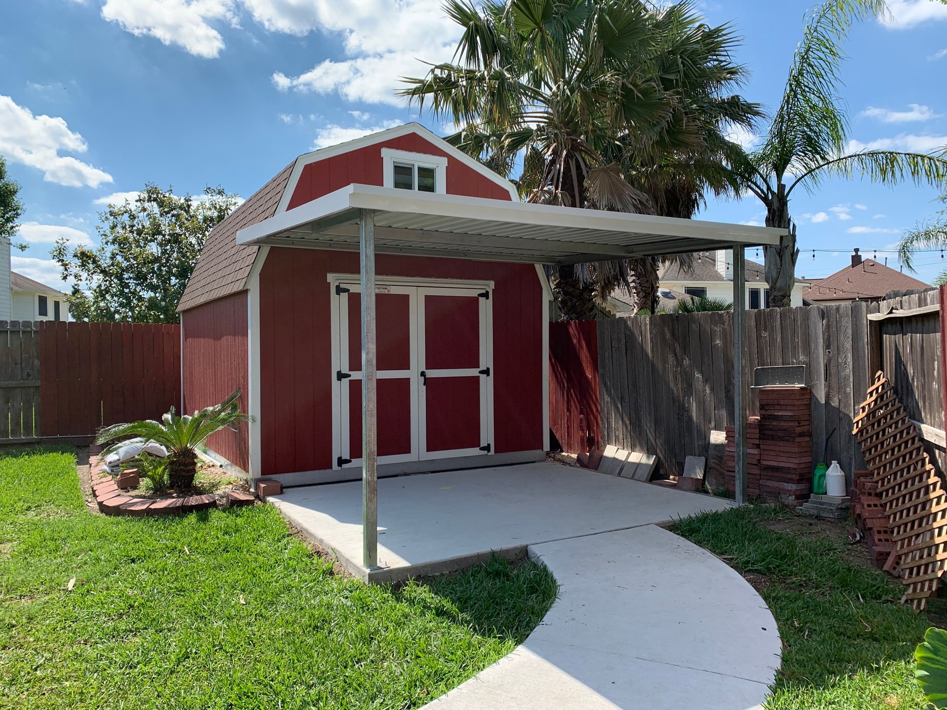 Red shed with a covered area, in a backyard. Concrete walkway, wooden fence, and grass.