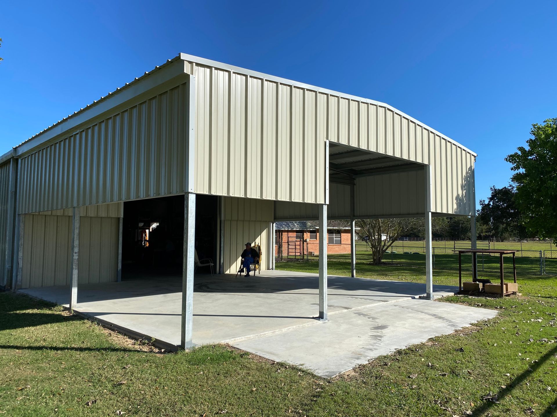 Metal barn with open-air carport on a concrete pad, on a grassy lawn under a clear blue sky.