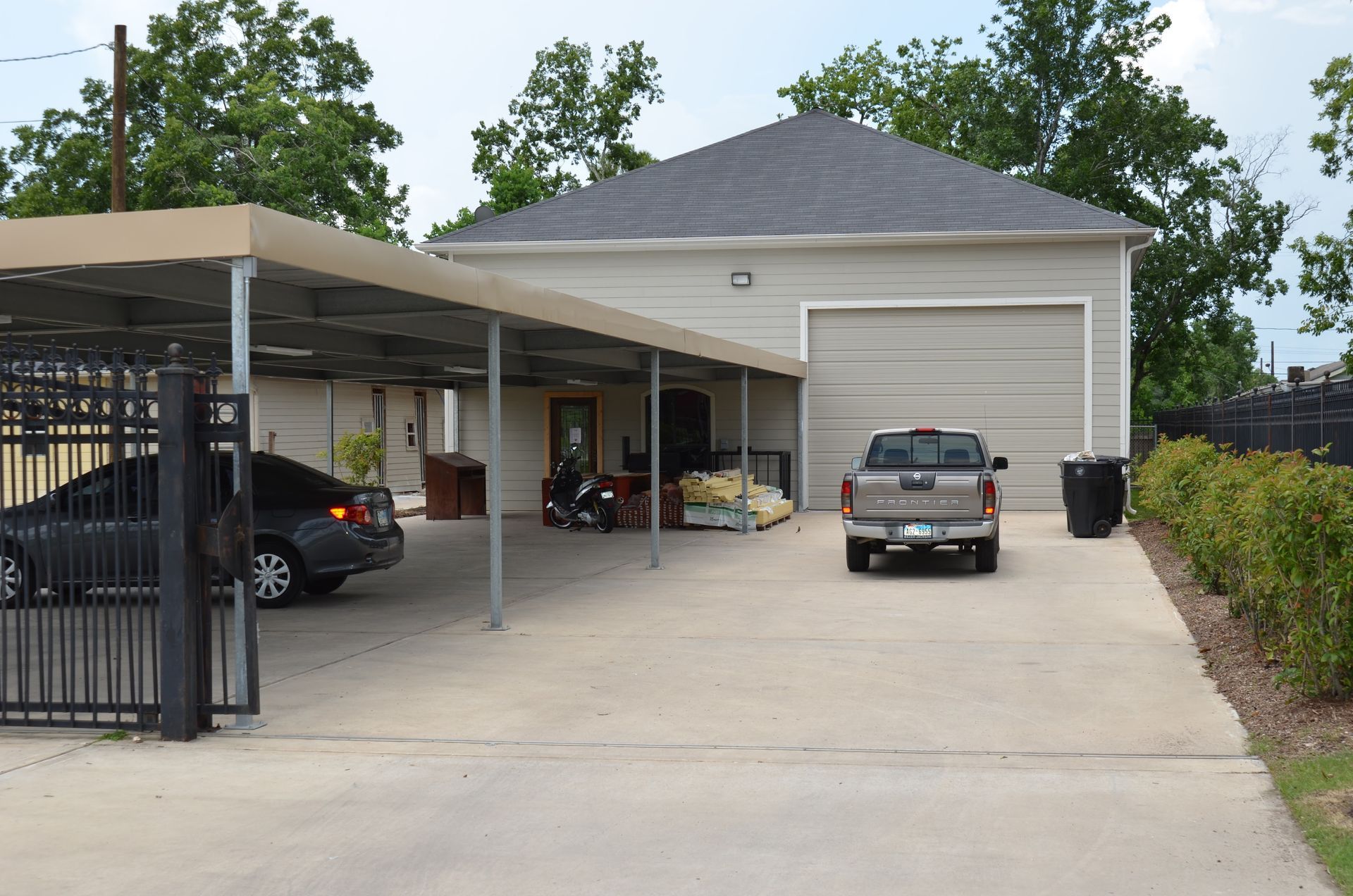 A driveway with a car parked under a metal carport, a pickup truck, and a garage in front of a building.