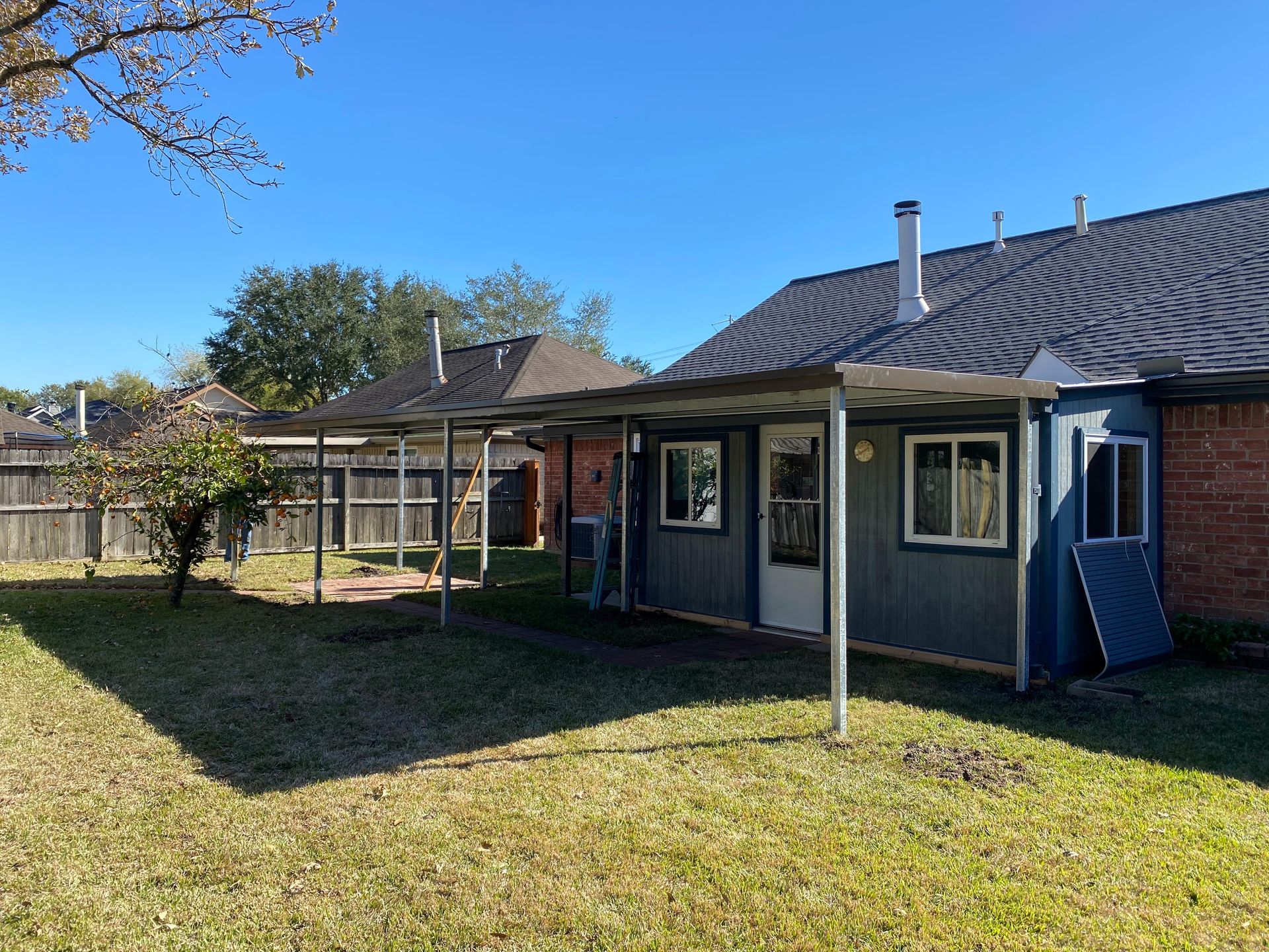 Backyard with a blue house, attached metal canopy, and green grass on a sunny day.
