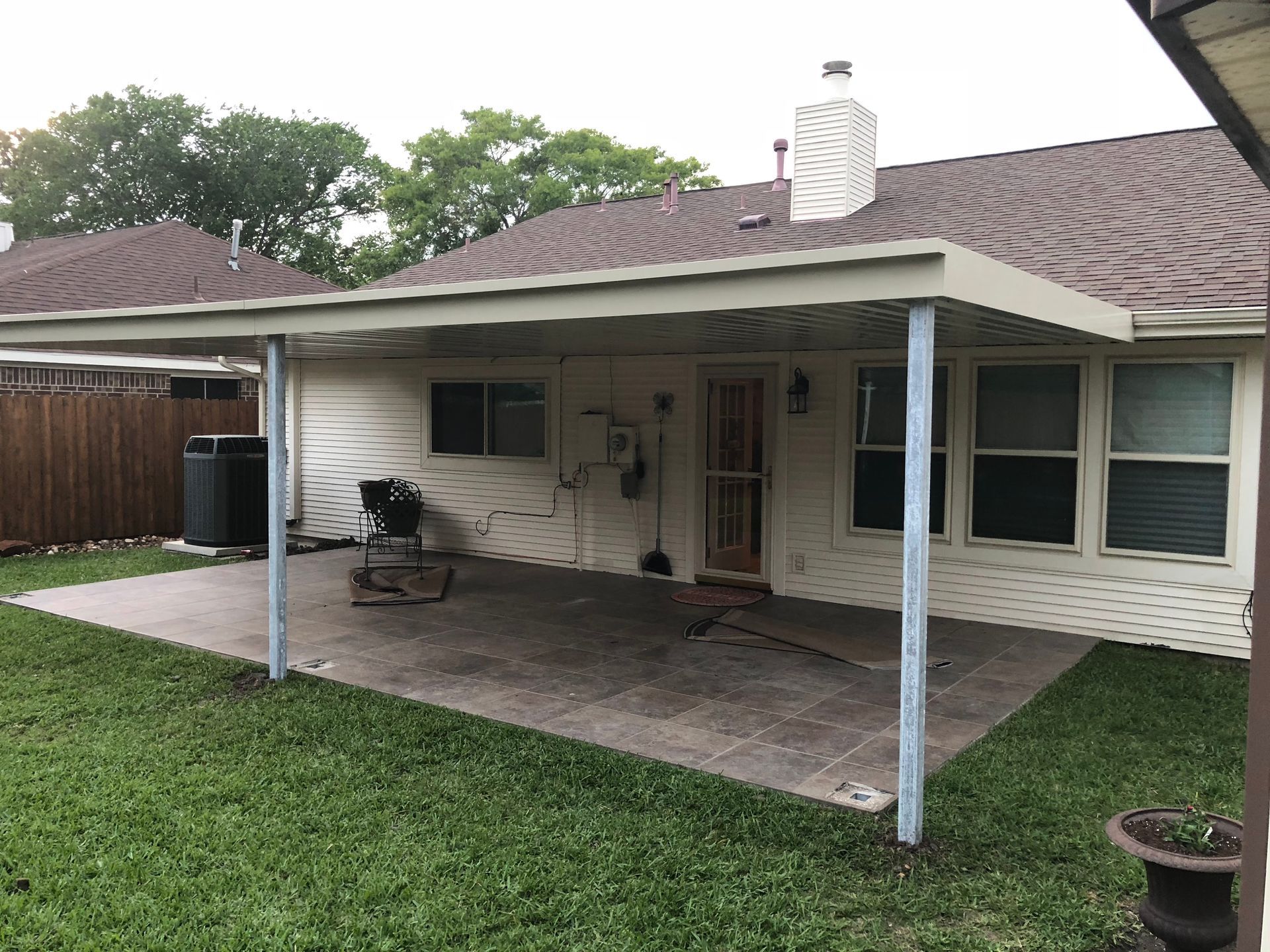 Patio with tan awning, concrete slab, and green grass. House and trees are in the background.