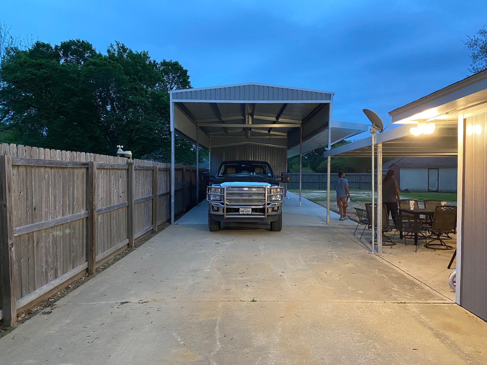 Truck parked under a carport at dusk.  Two people are near a building on the right.  A wooden fence is on the left.