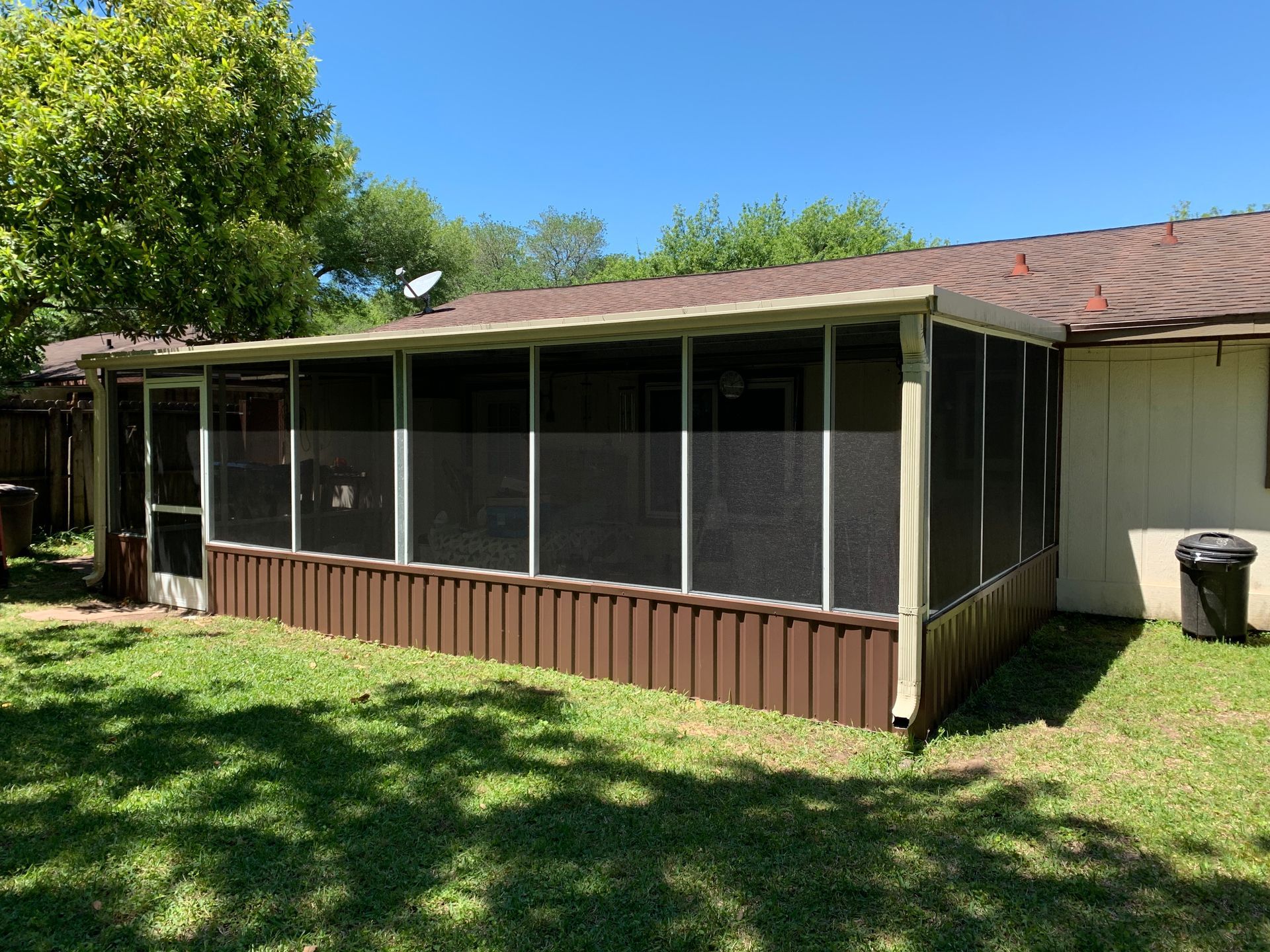 Screened porch with brown siding, attached to a house with a brown roof, on a sunny day.