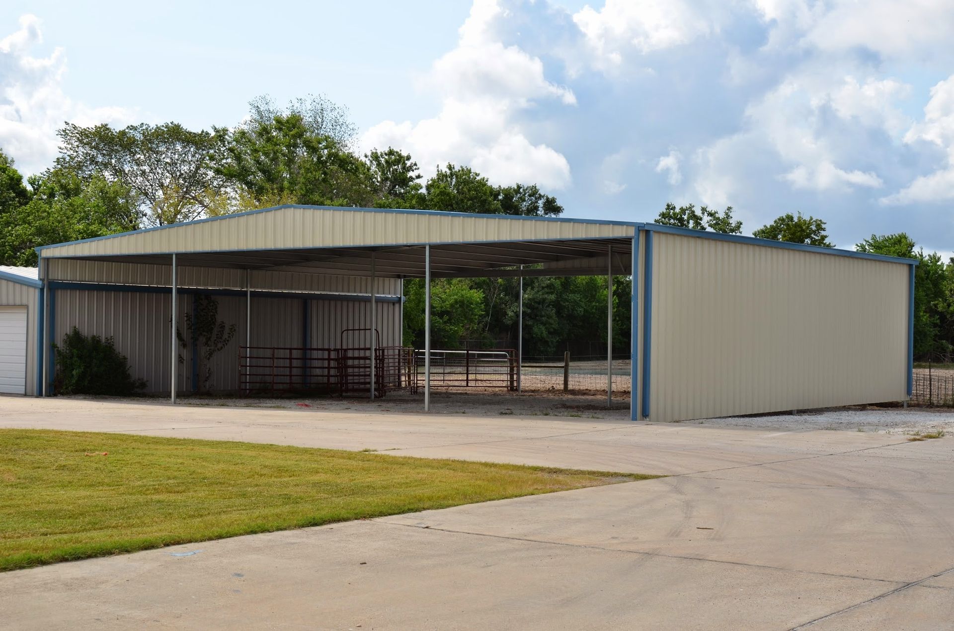 Metal shed with open sides and a gravel driveway in front. Green grass and trees visible.